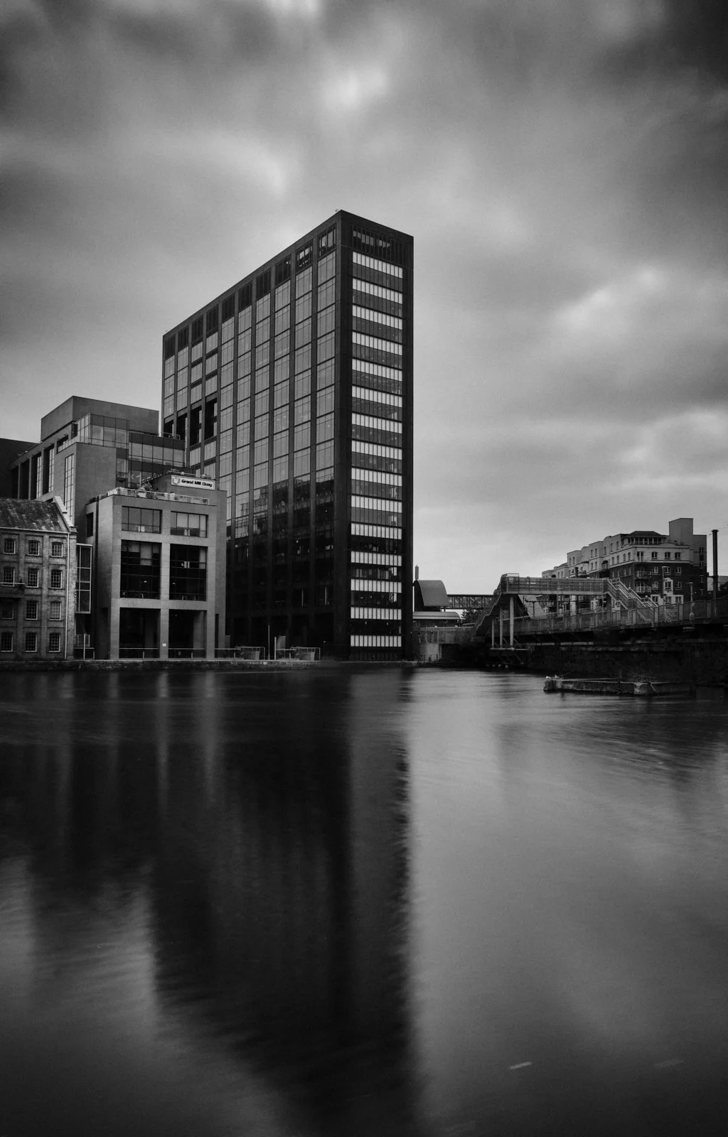 Black and white photo of a cityscape with modern high-rise buildings next to a river, cloudy sky overhead.