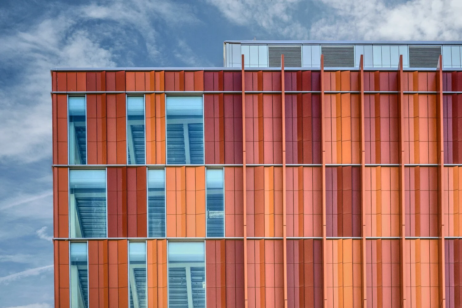 Close-up of a modern building facade with vertical orange panels, large glass windows, and a partial view of the sky with clouds.
