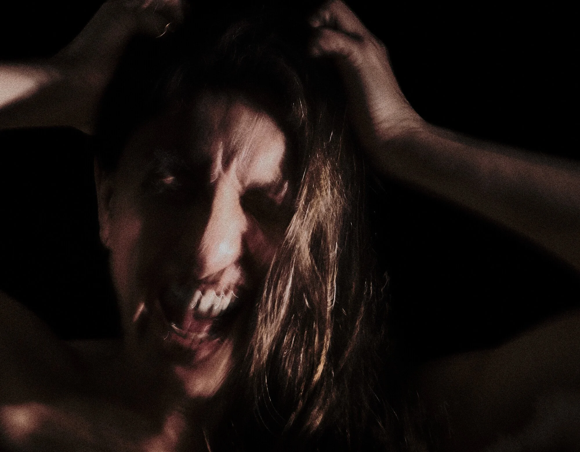 A woman with long hair biting her lip and has an intense expression, holding her head with both hands, against a dark background.