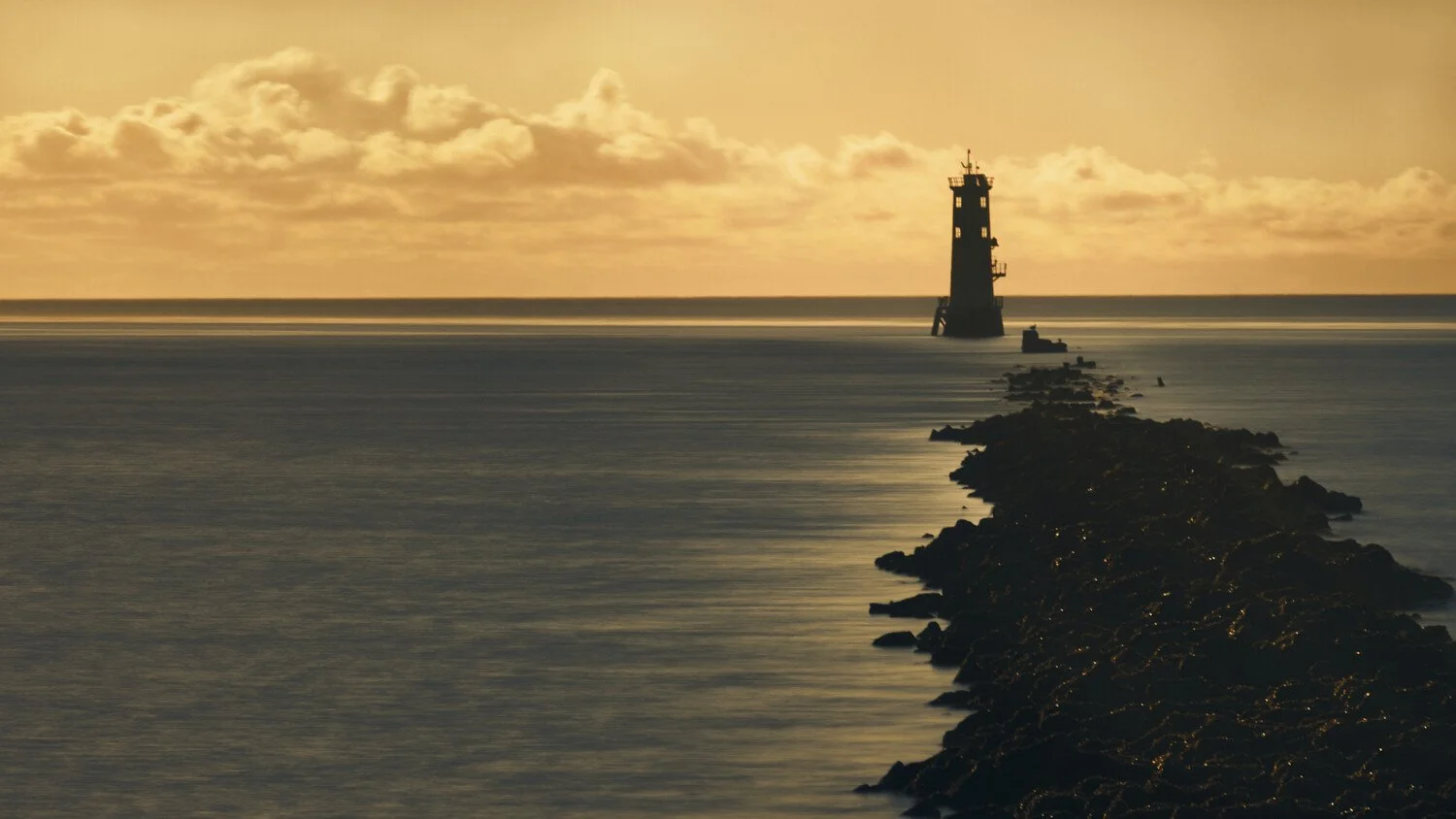 A lighthouse is located at the end of a rocky jetty extending into a calm body of water at sunset, with a partly cloudy sky in the background.