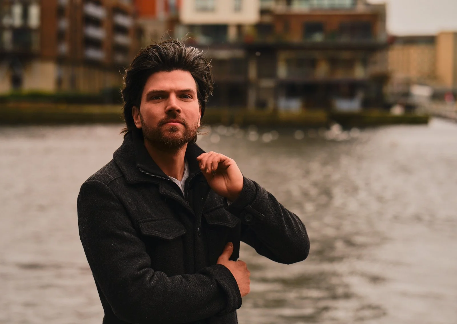 A man with dark hair and a beard standing outdoors near water, with modern buildings in the background.