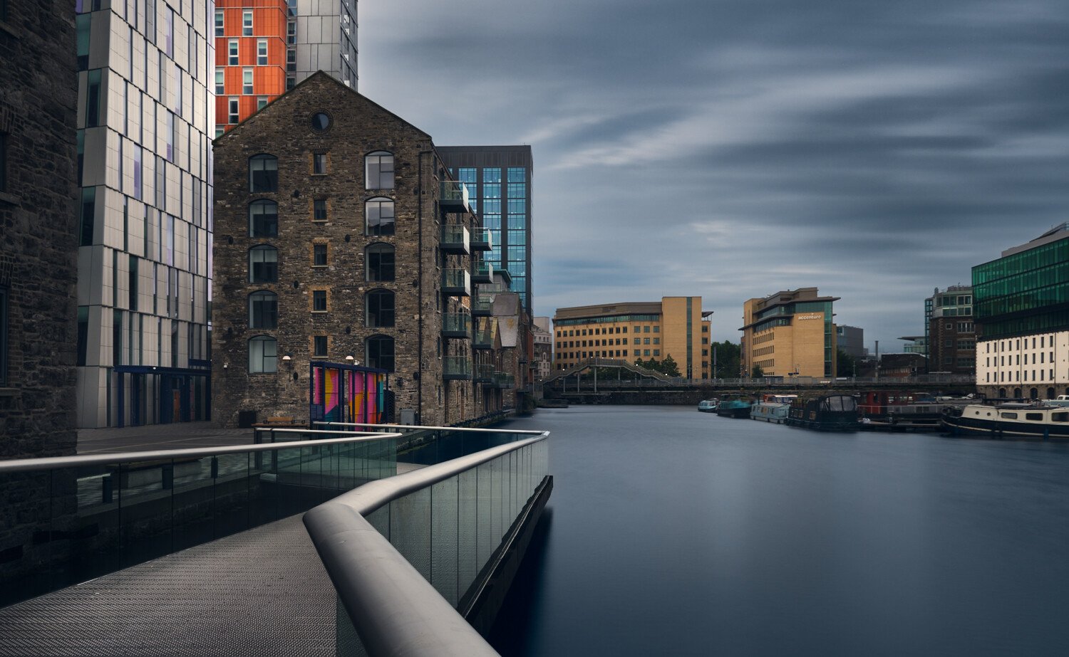 Cityscape with modern buildings along a waterway, including an older stone building, under a cloudy sky.