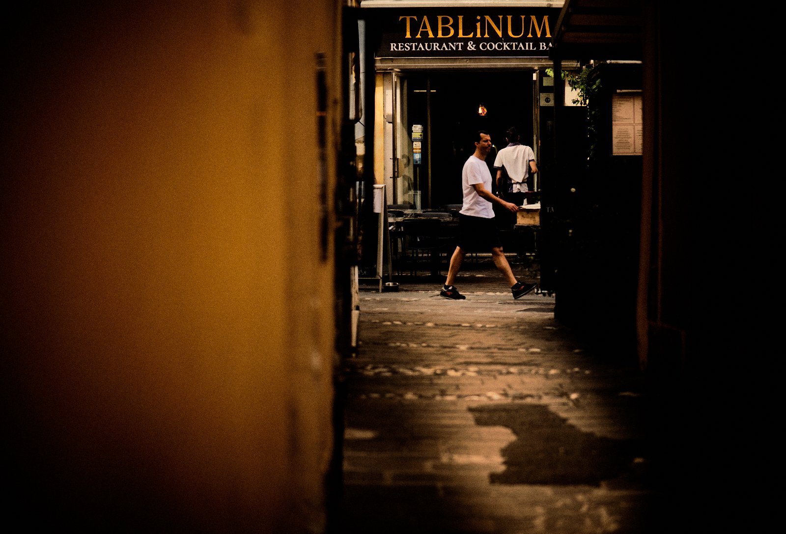 Restaurant terrace with a glowing golden sign seen through a dark passageway in Desenzano del Garda, a waiter and a passerby crossing paths — Provincia Malinconica by Tom Imio