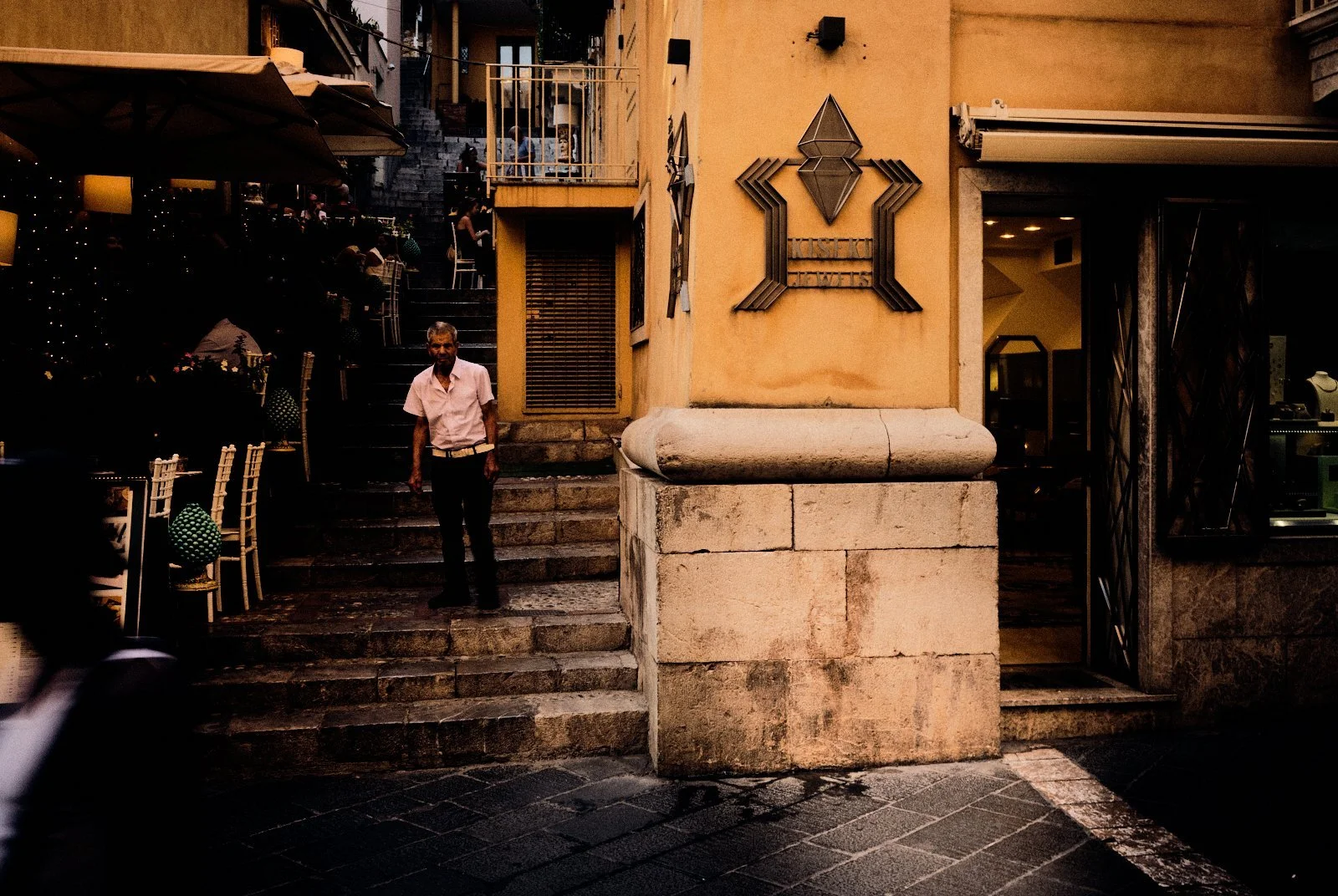 Elderly man standing on worn stone steps between a lamplit restaurant terrace and a jewellery shop in Taormina, Sicily — Provincia Malinconica by Tom Imio