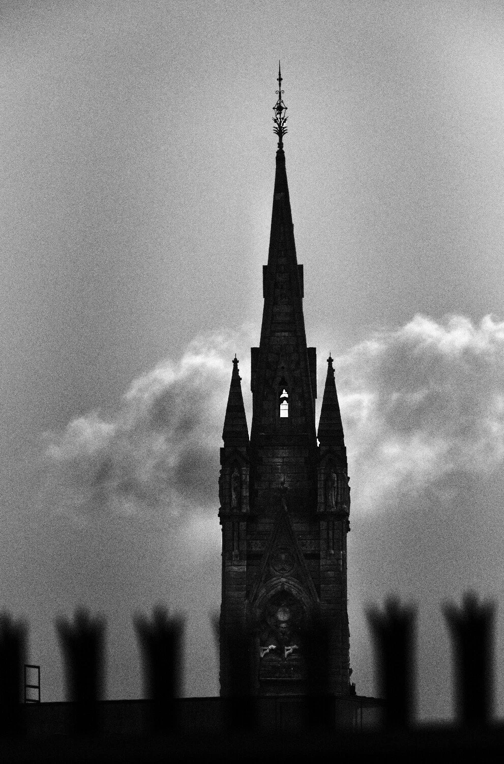 Black and white photo of a tall, Gothic-style church steeple with a spire and decorative details, seen against a cloudy sky.