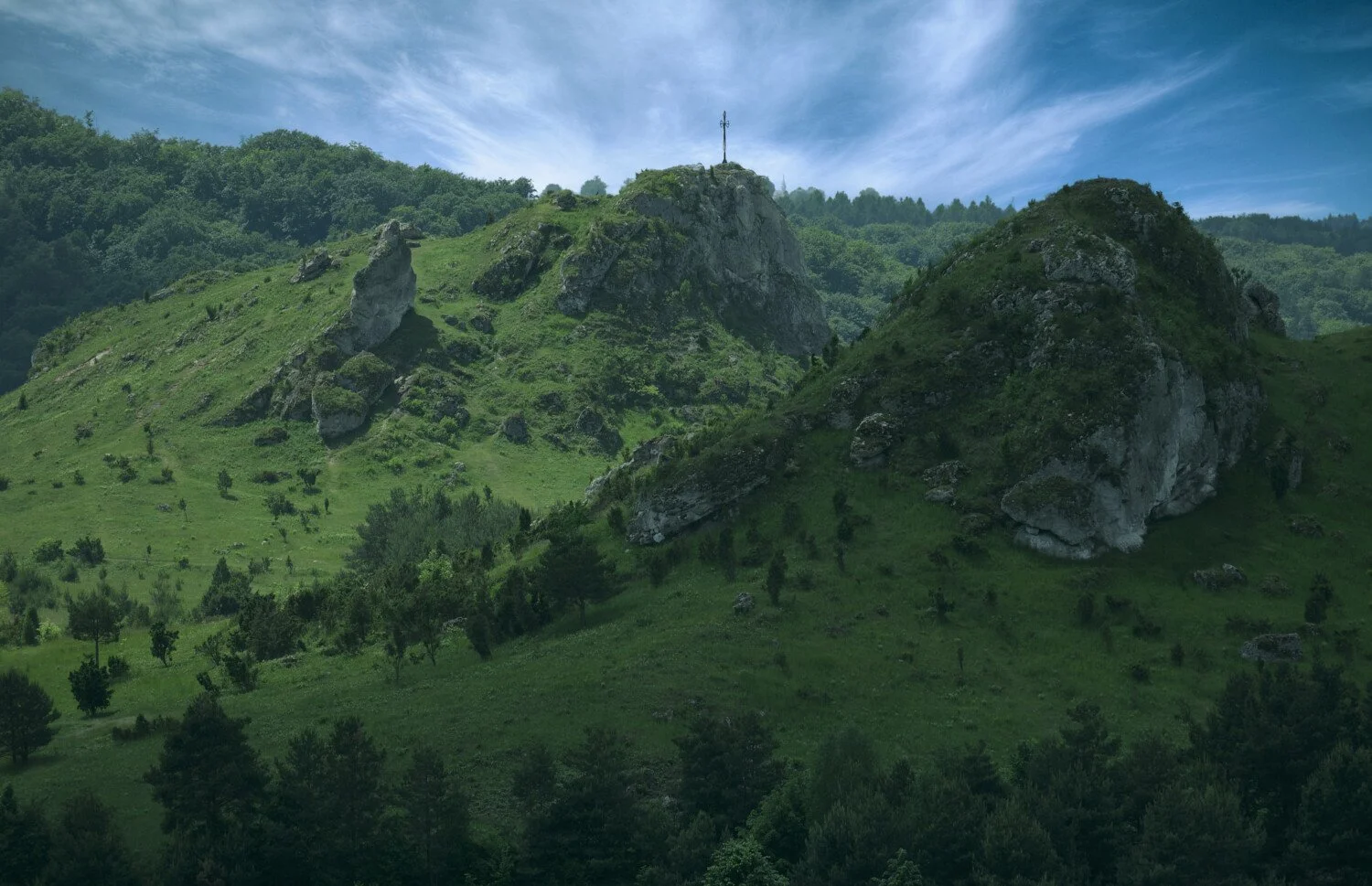 Green hillside with large rocks and a cross on top of the hill under a partly cloudy sky.