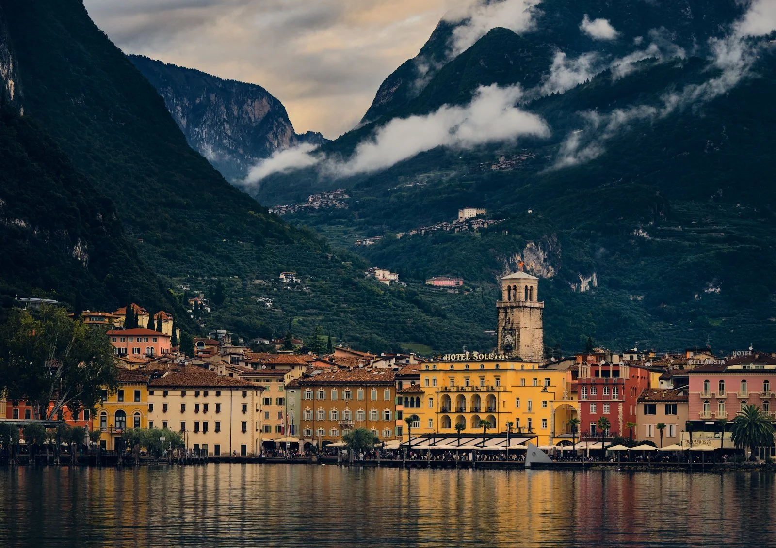 Colourful buildings along a waterfront with a mountain landscape and clouds in the background. Riva del Garda. Italy