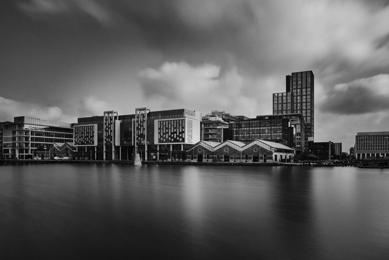 Black and white photo of modern buildings along a waterfront with cloudy sky.