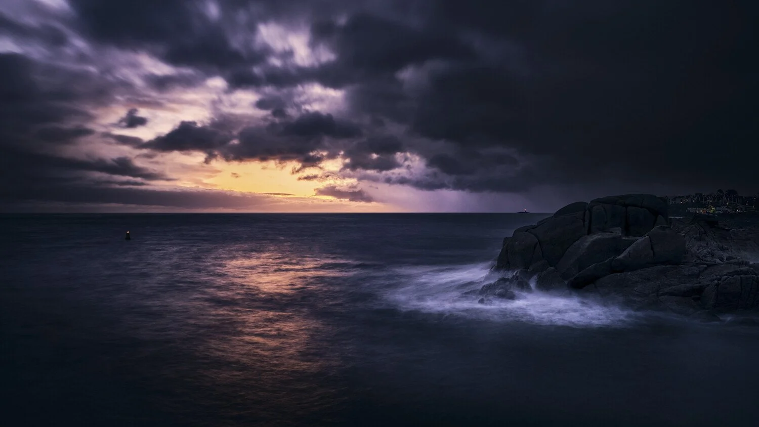Dark ocean scene at sunset with stormy clouds overhead, large rocks on the right side, and a small lighthouse in the distance.