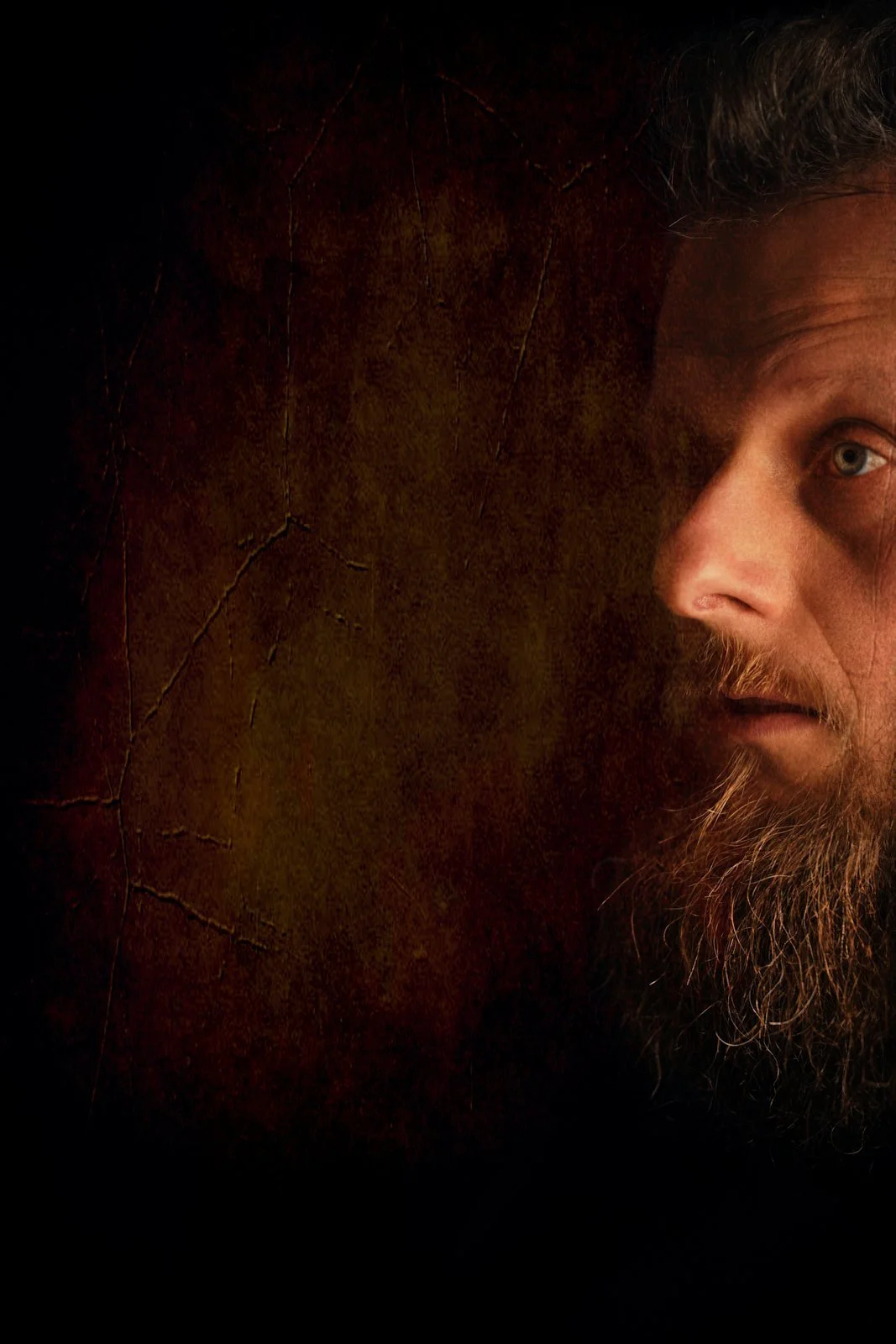 A close-up of a bearded man with hair, looking to the side, with a dark textured background.