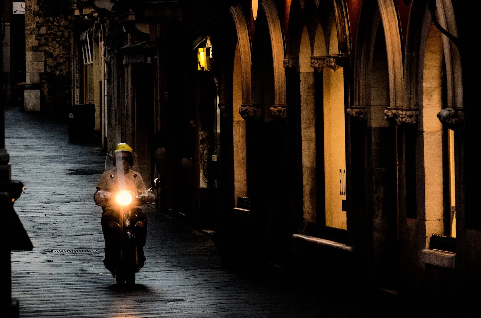 Scooter with a glowing headlamp riding past Gothic arcades on an empty street at dawn in Taormina, Sicily — Provincia Malinconica by Tom Imio