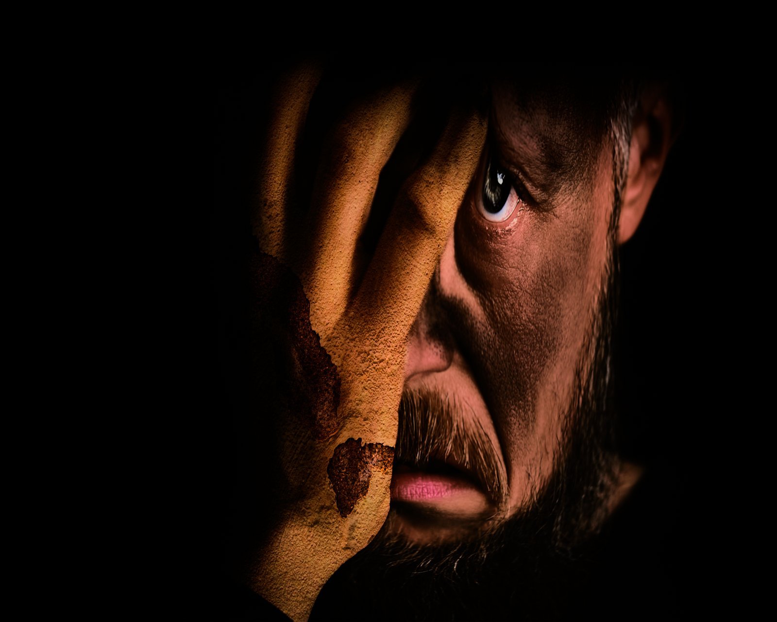 A close-up of a man with intense eyes, holding his face with one hand, partially obscuring his features in low light.