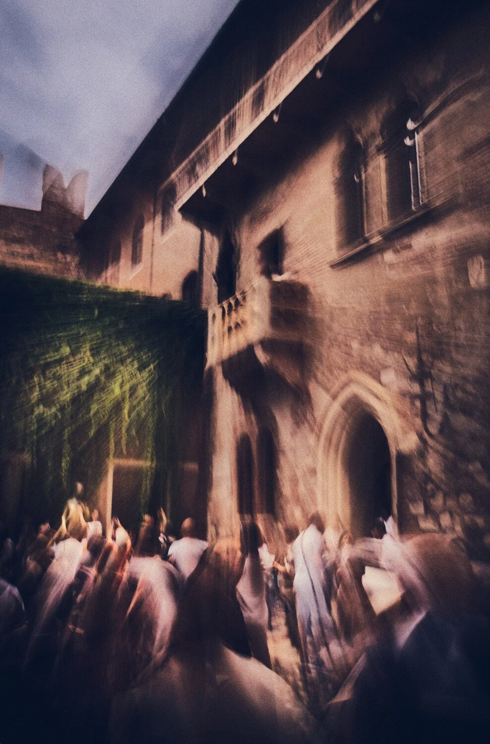 A nighttime scene of a historic brick building with arched windows and a tower, with a crowd of people gathered in the foreground. The photo has a motion blur effect.