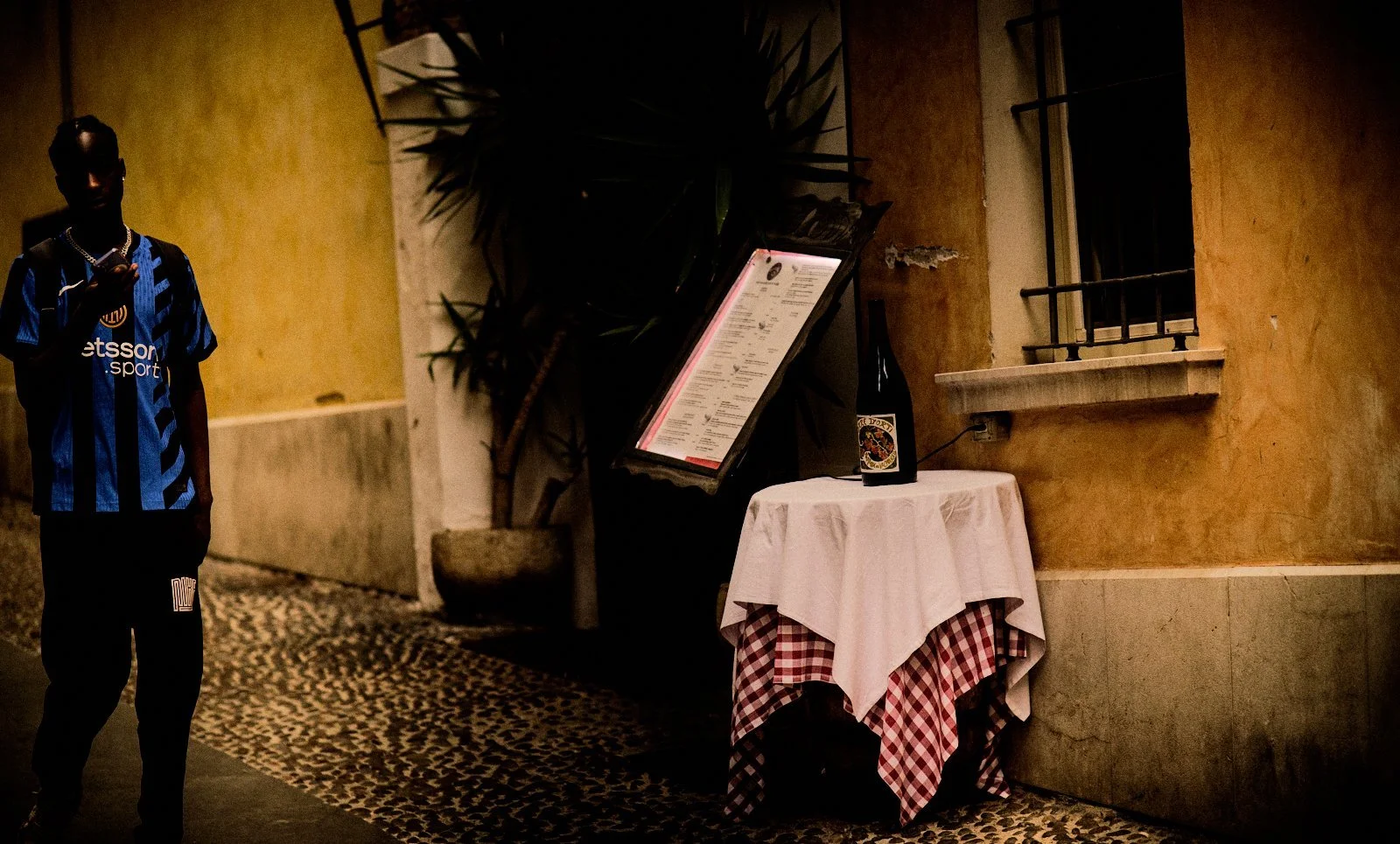 Young man in a football shirt emerging from shadow beside a trattoria table with a checked tablecloth and wine in Desenzano del Garda — Provincia Malinconica by Tom Imio