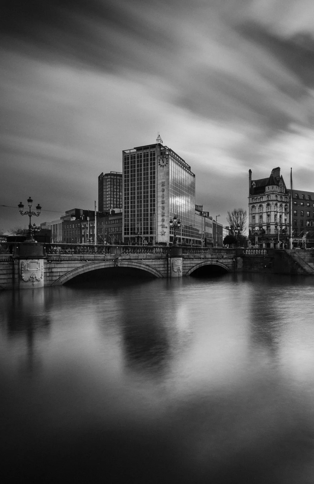 Black and white photo of a cityscape with a river in the foreground, featuring a bridge and tall buildings, including one with the sign 'Heueller X' and a clock, under a sky with moving clouds.