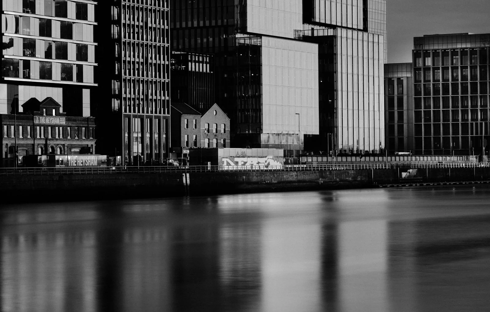 Black and white photo of modern city buildings along a river or canal, with reflections on the water and graffiti on a wall in the foreground.