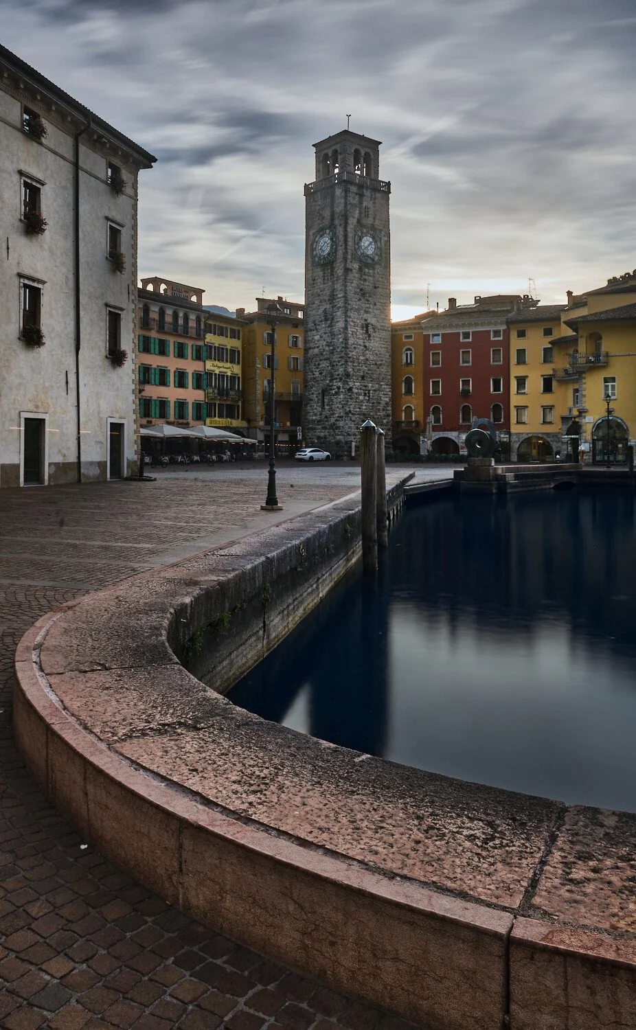 Historic town square with a stone clock tower, colorful buildings, a water fountain, and cloudy sky.