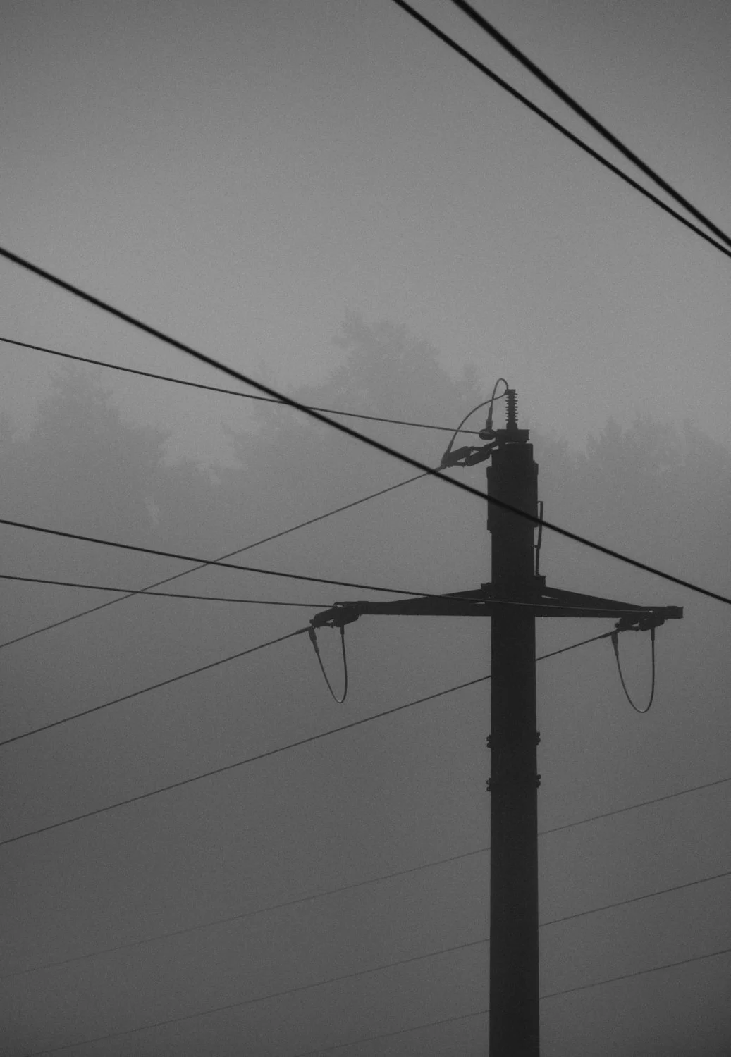 A black and white image of a utility pole with electrical wires against a cloudy sky.