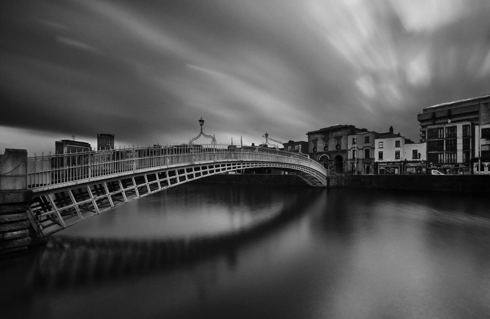 Black and white photo of a bridge over a river, with buildings in the background and a cloudy sky.
