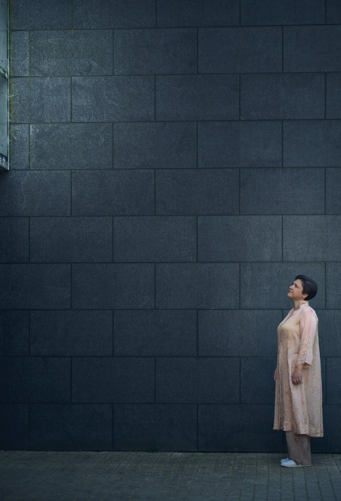 Woman in a pale pink outfit standing alone before a vast dark tiled wall, looking upward