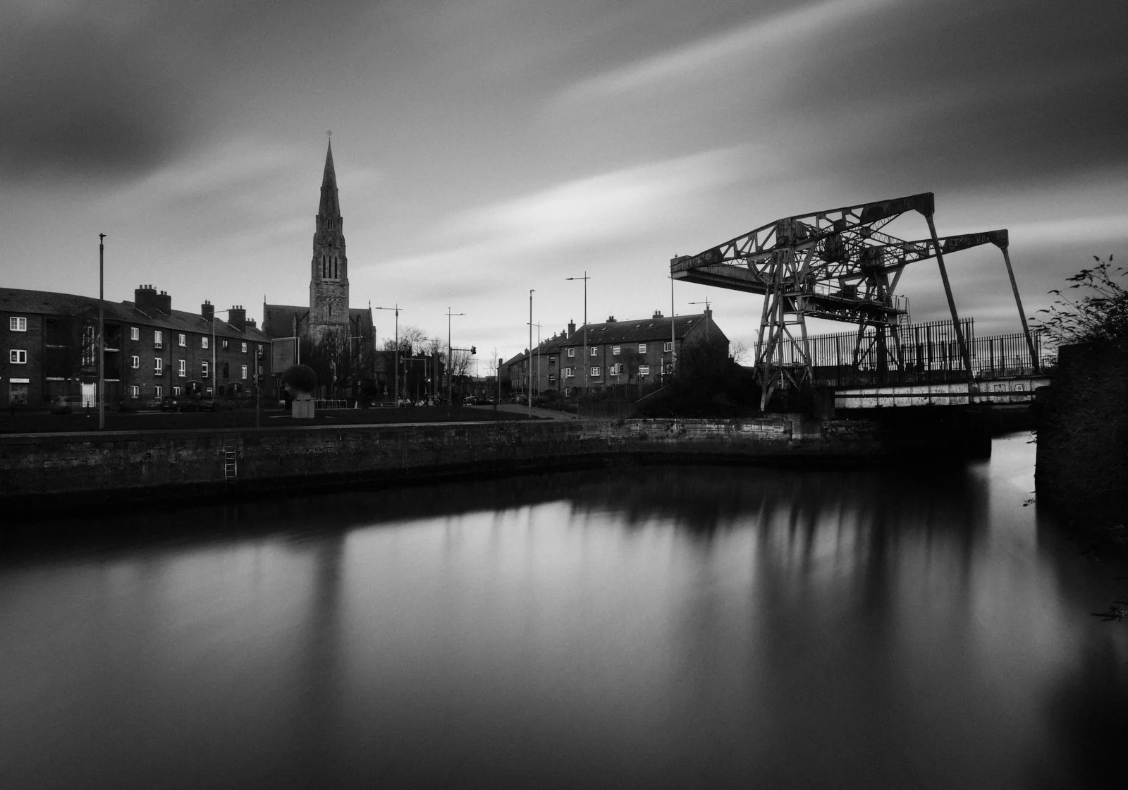 Black and white photo of a river with reflections, a church with a tall spire, residential buildings, and an industrial crane on the riverbank under a cloudy sky.
