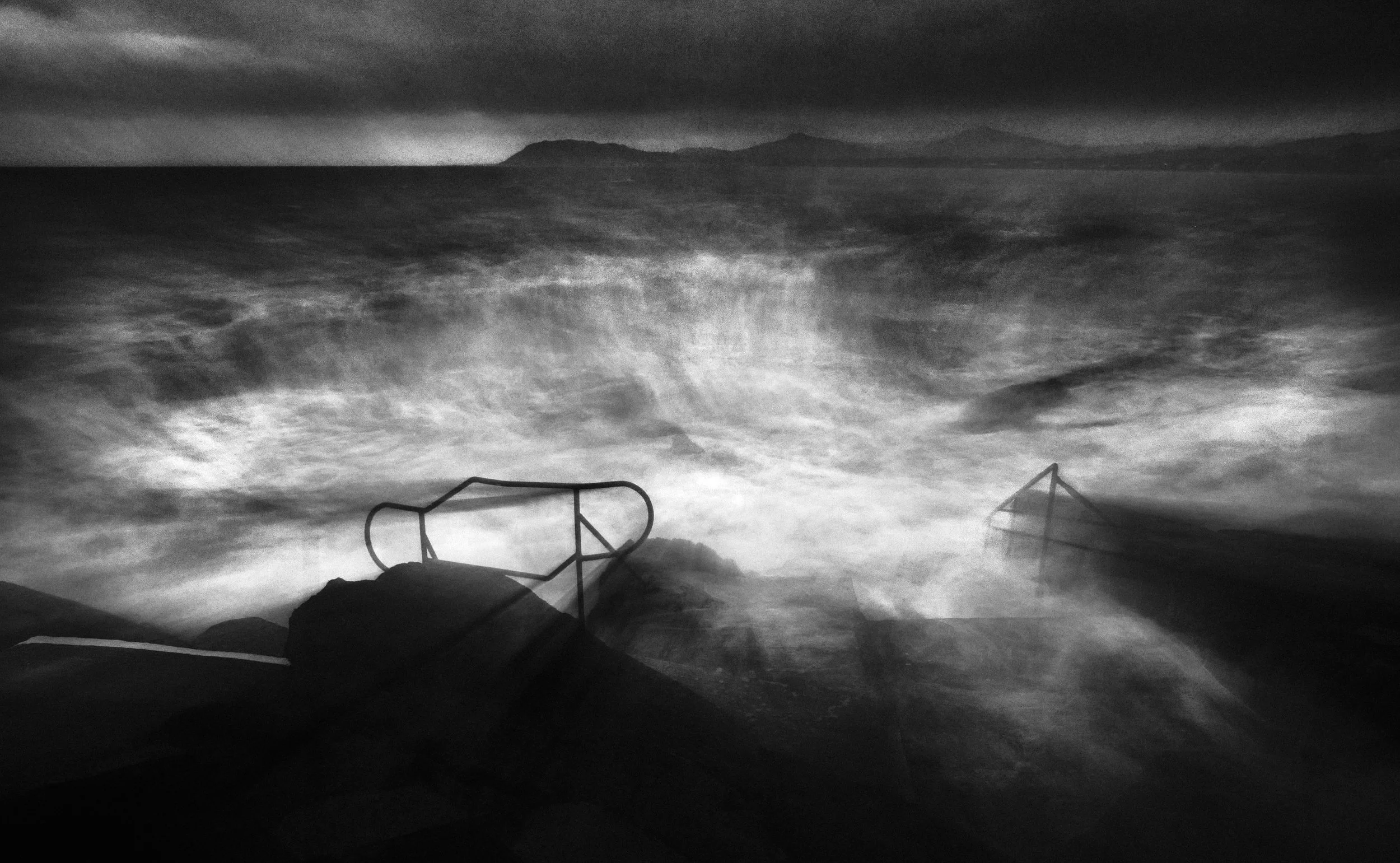 Black and white photograph of a cloudy sky over a landscape with a structure in the foreground and distant mountains in the background.