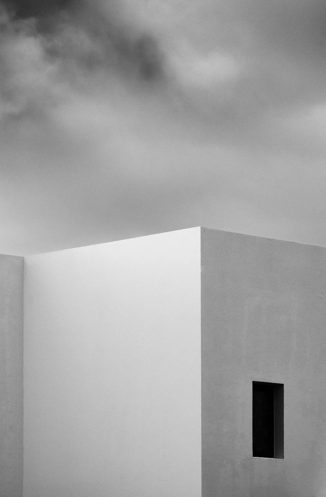 Black and white photo of a modern white building with smooth walls, a small rectangular window, and a cloudy sky above.