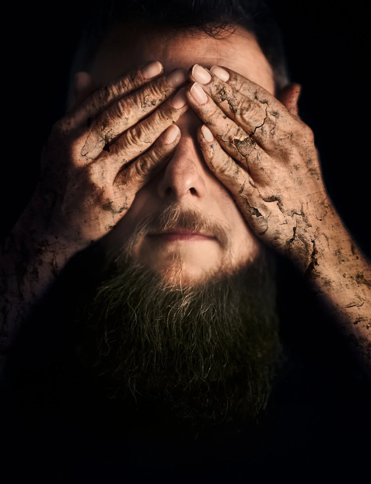 A man with a long beard covers his eyes with his muddy hands.