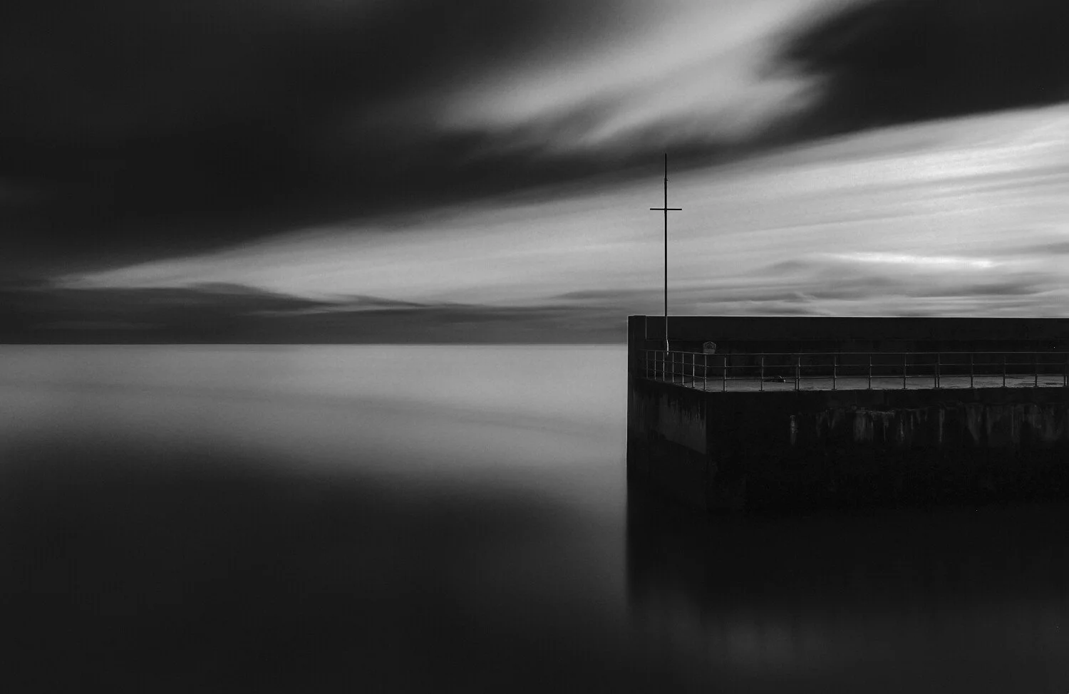 Black and white photo of a pier extending into calm water with a metal railing and a tall pole, under a dark, cloudy sky.