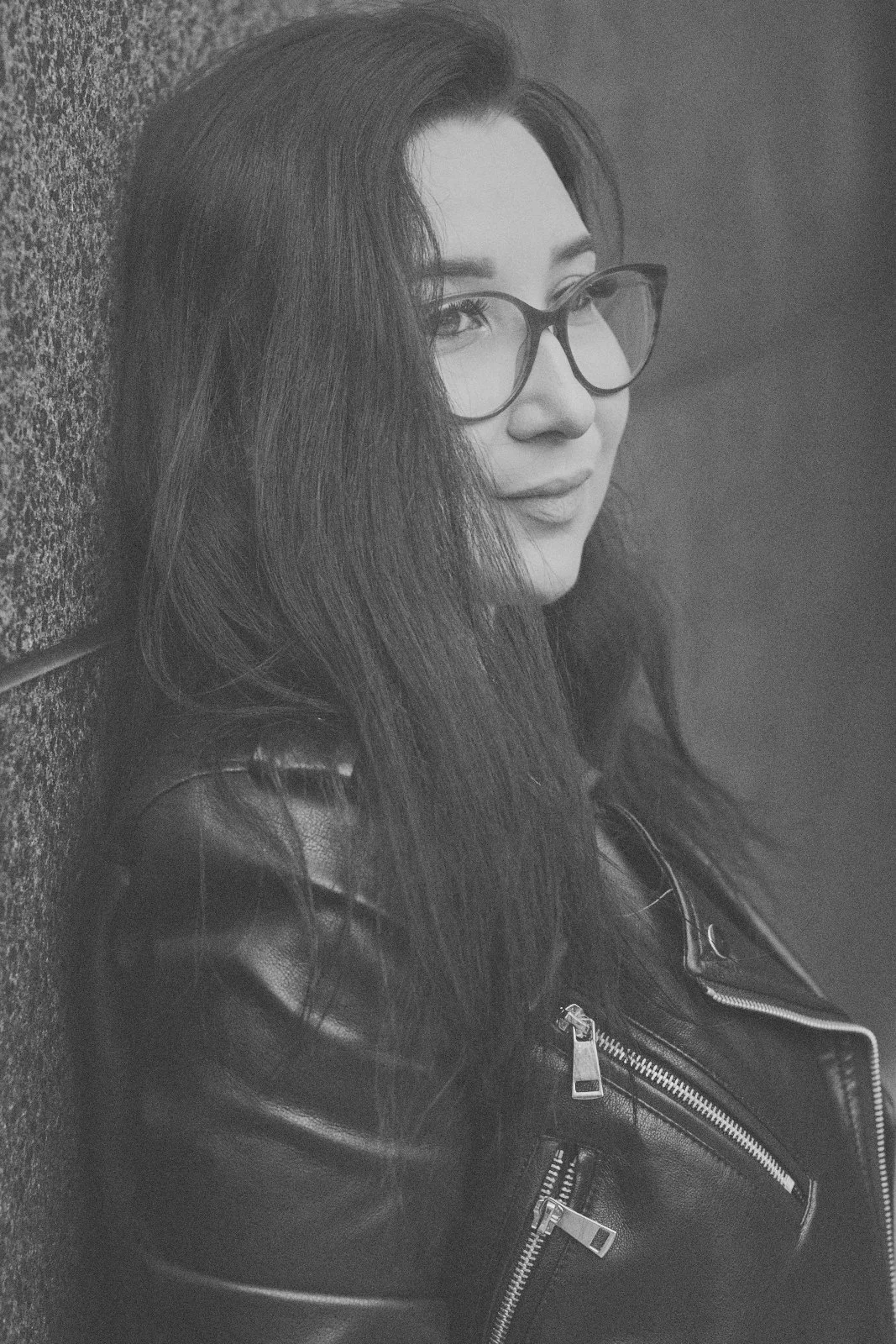Black and white portrait of a woman with long dark hair and glasses in a leather jacket leaning against a stone wall