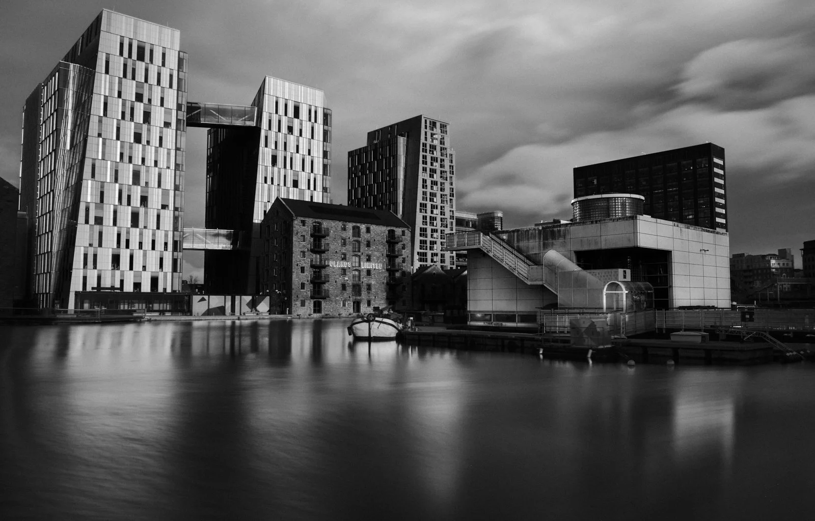 Black and white photo of modern city buildings along a waterfront with a boat docked close to the shore.