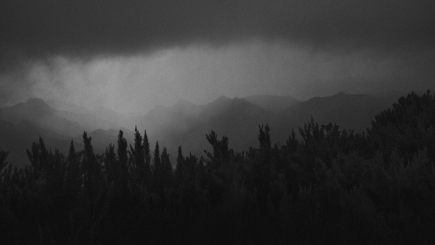 Black and white photograph of mountains with dark, stormy clouds overhead and a dense forest in the foreground.