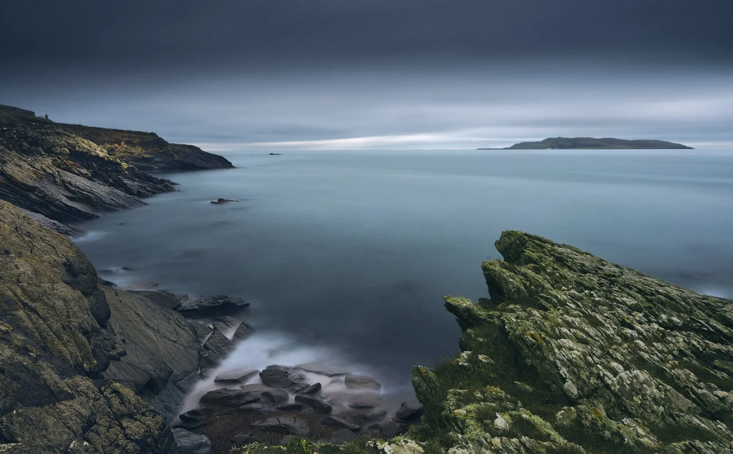 Calm ocean scene with rocky cliffs and an island in the distance under a cloudy sky.
