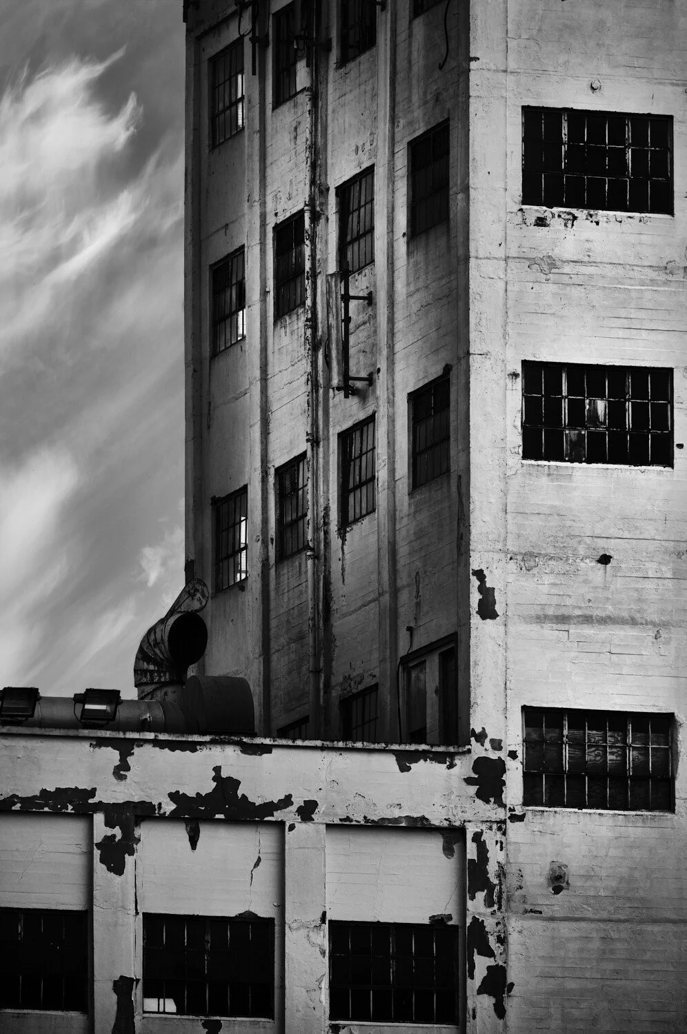 Black and white photo of a tall, abandoned industrial building with broken and boarded-up windows, peeling paint, and a cloudy sky in the background.