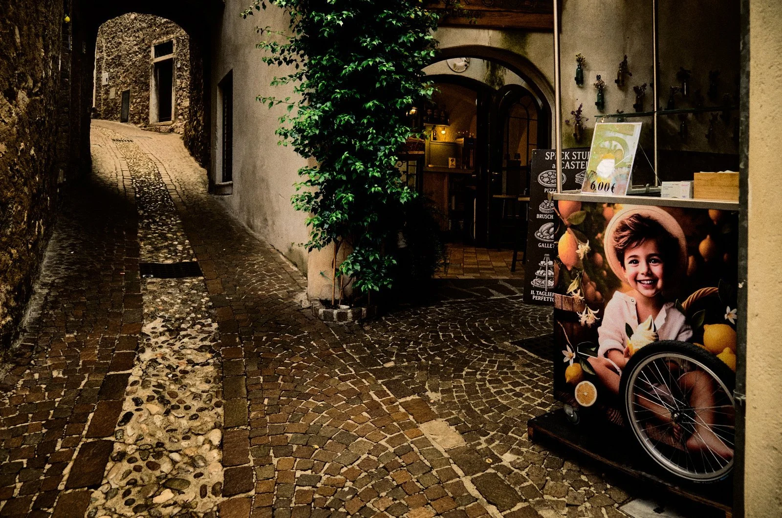 Weathered stone alley in Limone del Garda with a food cart advertising snacks beside real cobblestones and ivy — Provincia Malinconica by Tom Imio