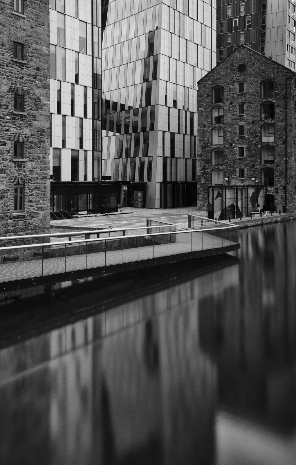 Black and white photo of modern glass buildings and old brick buildings next to a waterway with a bridge.