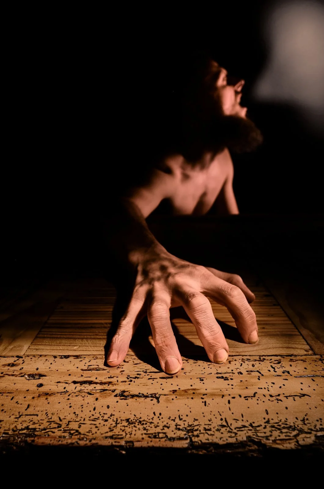 Close-up of a man with a beard reaching out and touching a wooden surface in darkness, with faint light highlighting his face and hand.