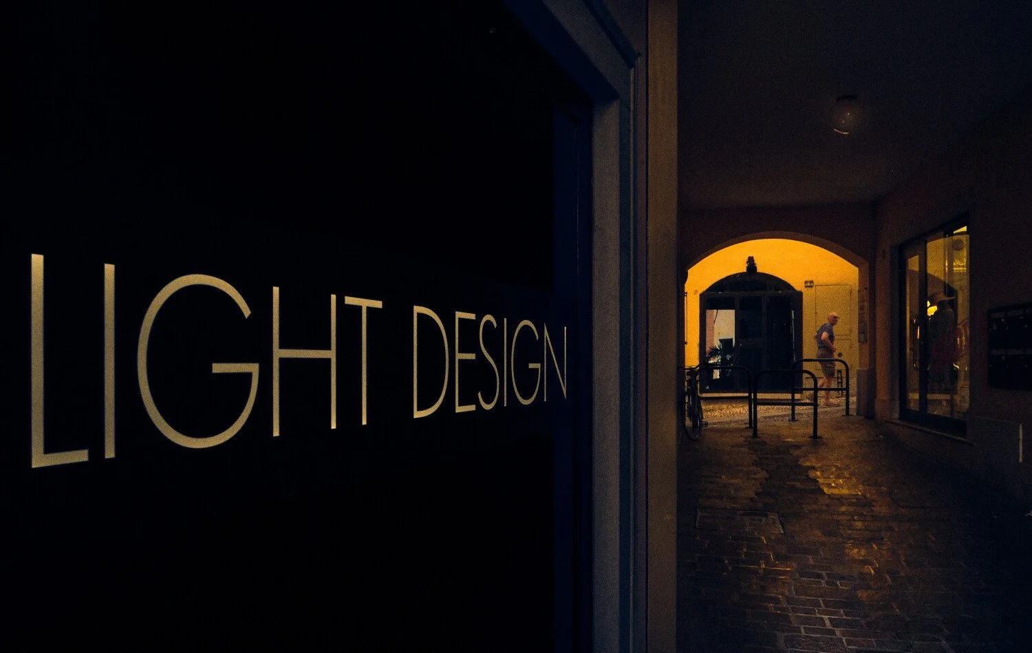 Illuminated "Light Design" shop sign in a dark passage, with a man standing in a warmly lit archway beyond