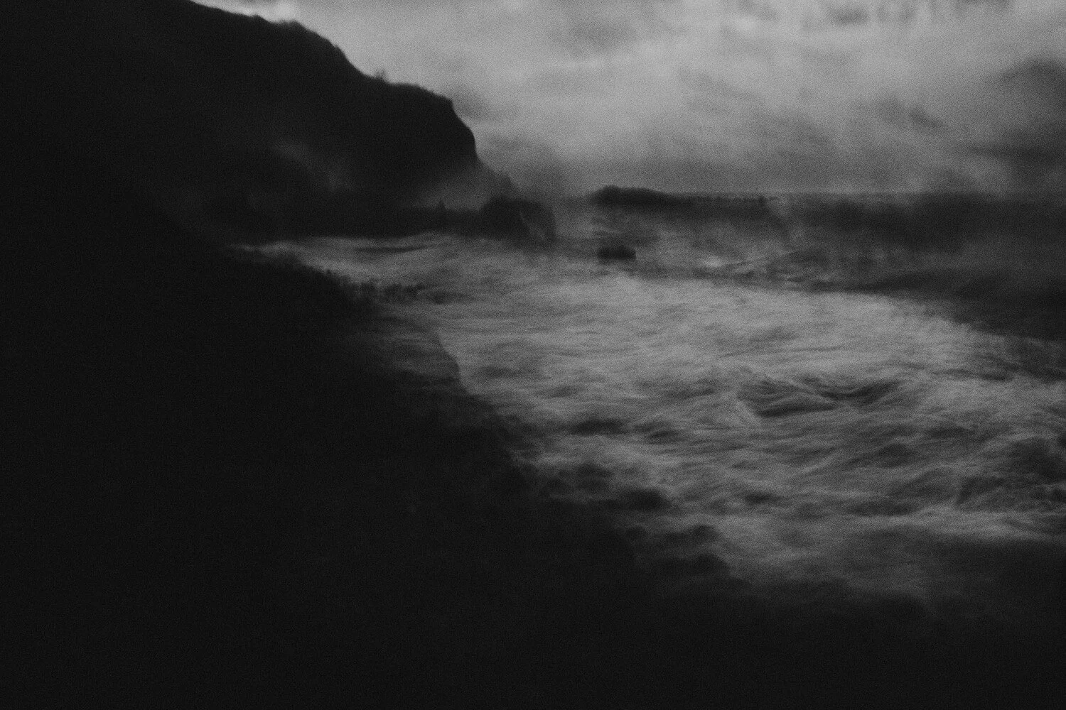 Black and white photograph of a rocky shoreline with waves crashing against the rocks under a cloudy sky.