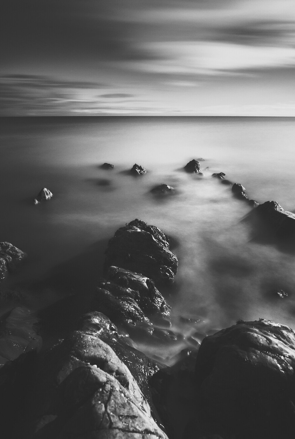 Black and white photo of rocks extending into water, with a calm sea and a cloudy sky in the background.