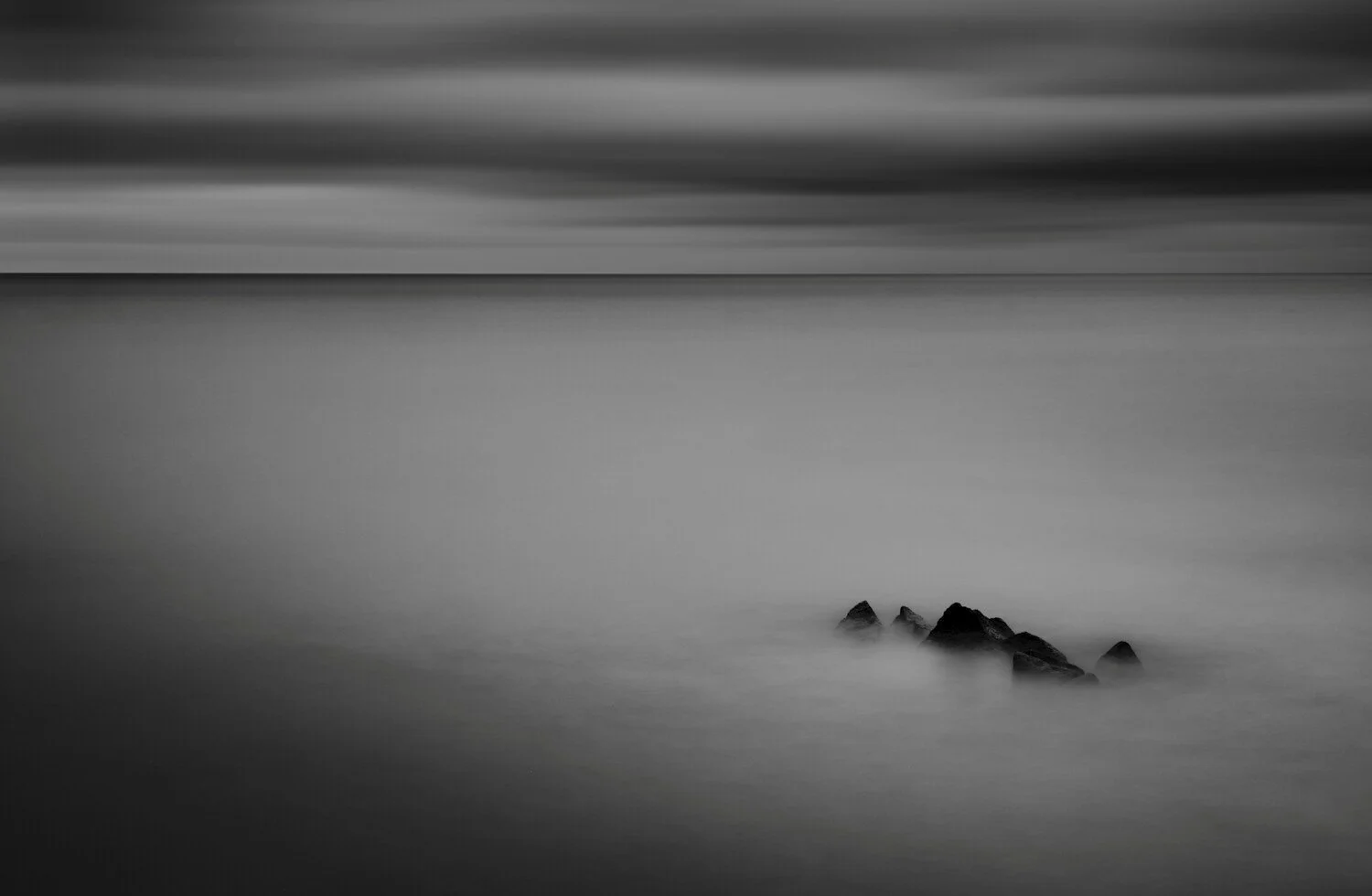 Black and white photo of a calm ocean with a small group of rocks partly submerged in water in the foreground. The sky and water appear smooth and misty.