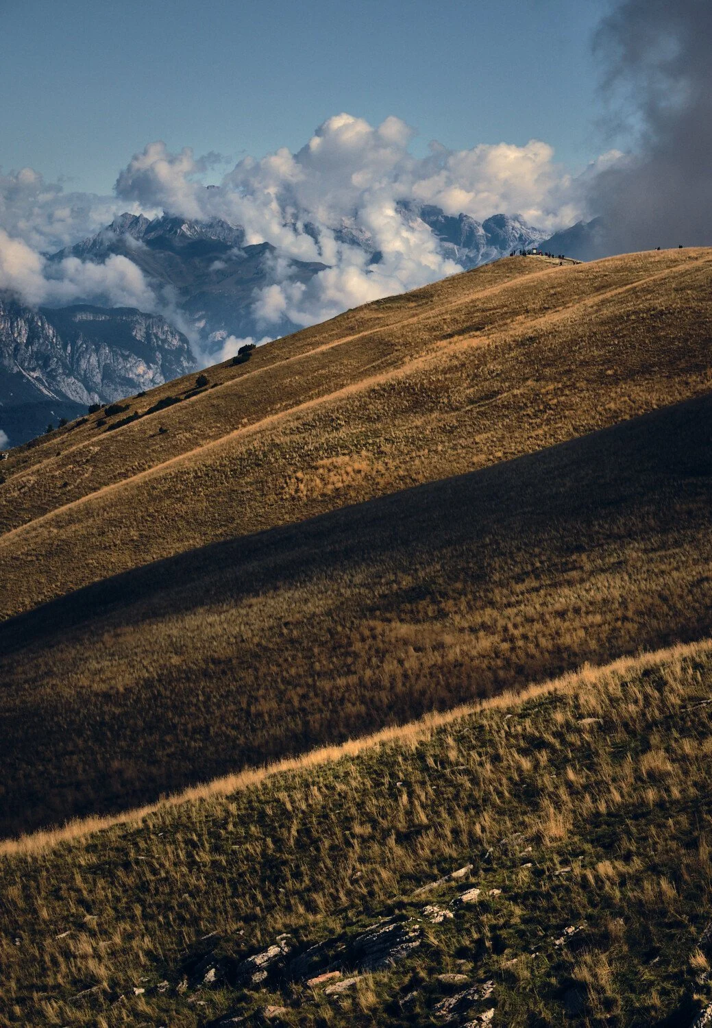 Rolling hills with dry grass, mountain peaks with snow and clouds in the background.