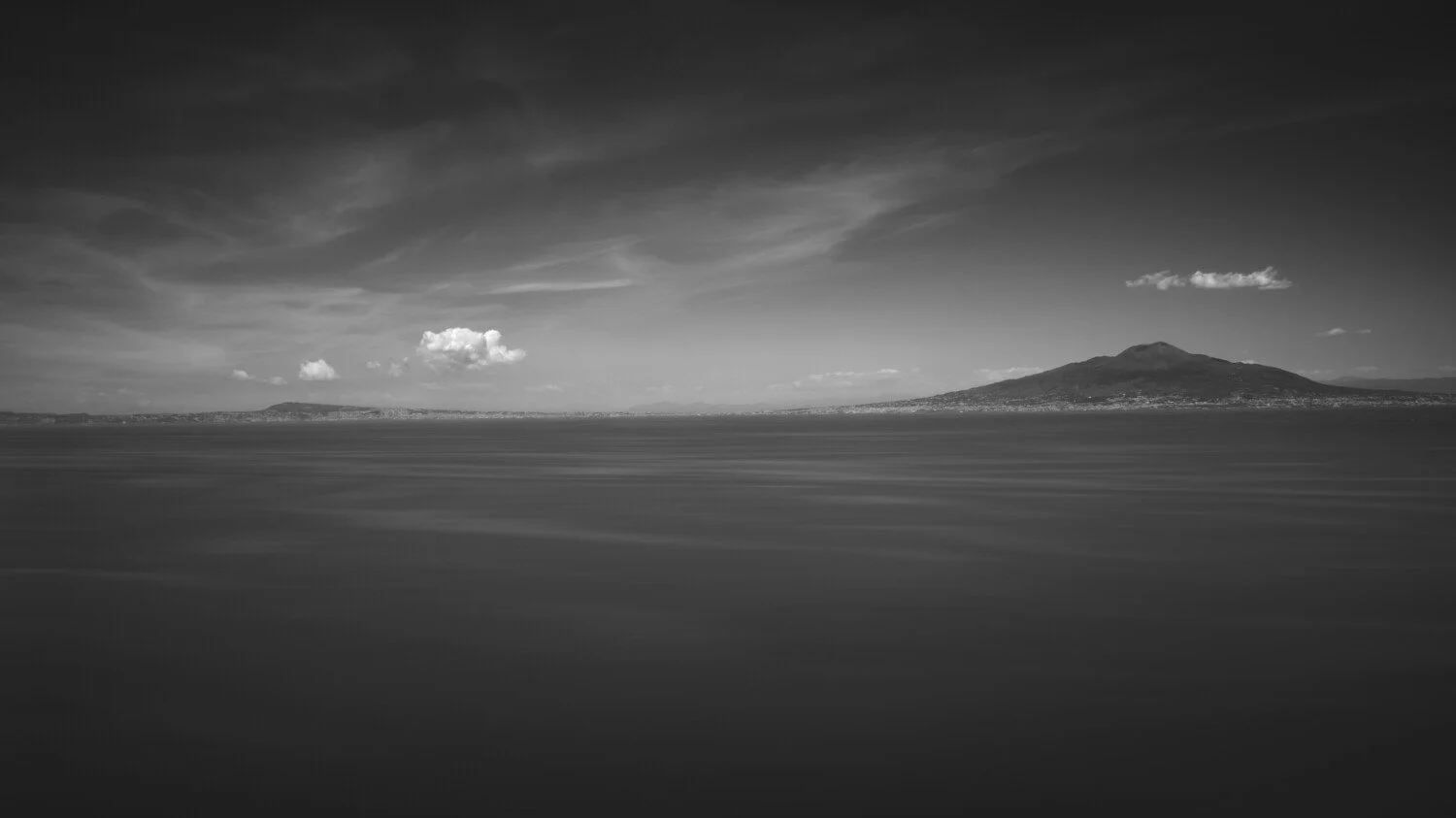 Black and white landscape photograph of a large body of water with distant hills or mountains under a cloudy sky.