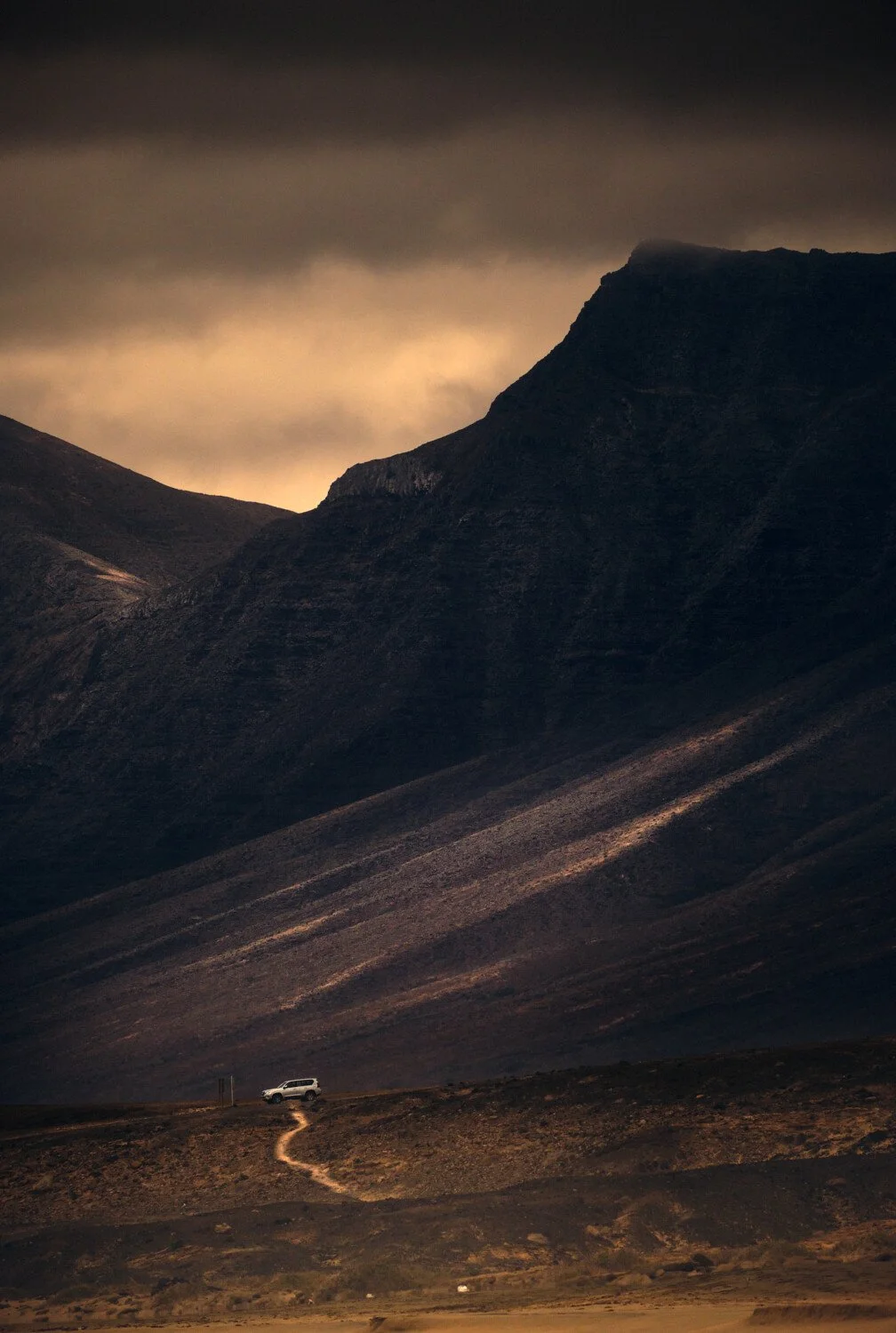 A white SUV parked on a dirt road at the base of dark, rocky mountains under a cloudy sky with a yellowish glow.