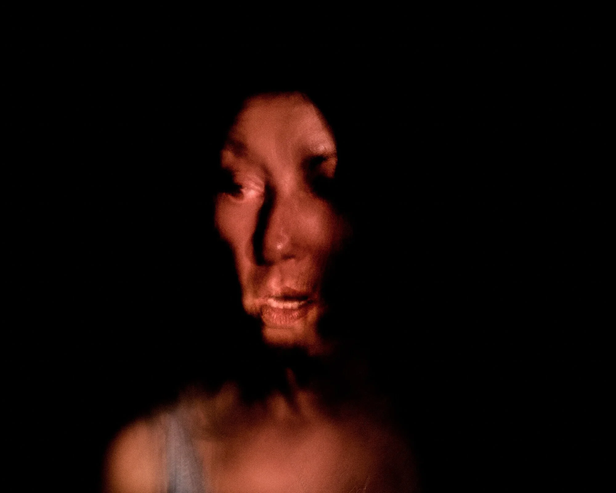 A woman with light skin, partially illuminated in darkness, making a mischievous or playful facial expression with a subtle smile and squinted eyes.