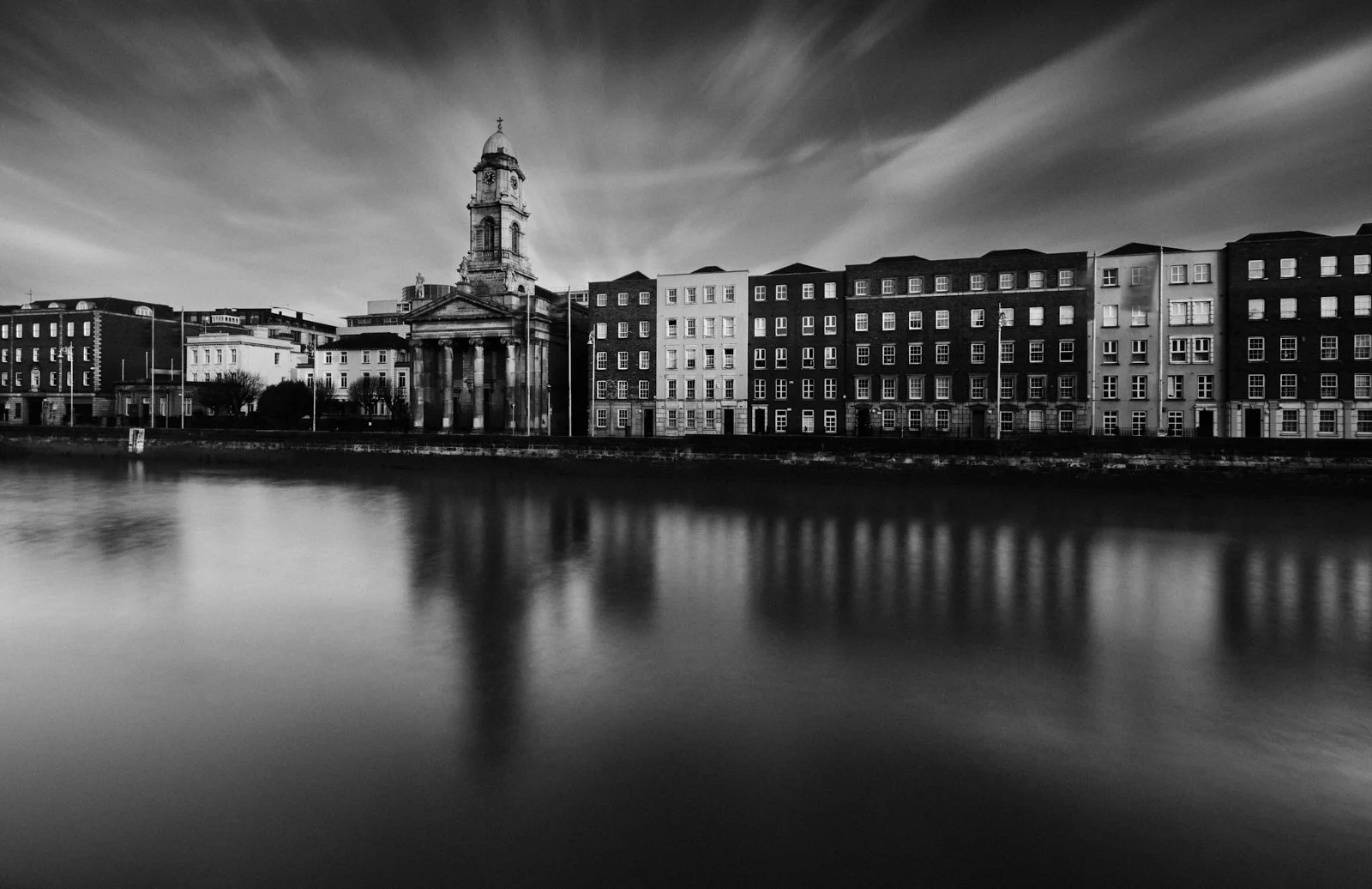 A black and white photo of a cityscape across a river, featuring a historic church with a tall bell tower and several multi-story buildings behind it.