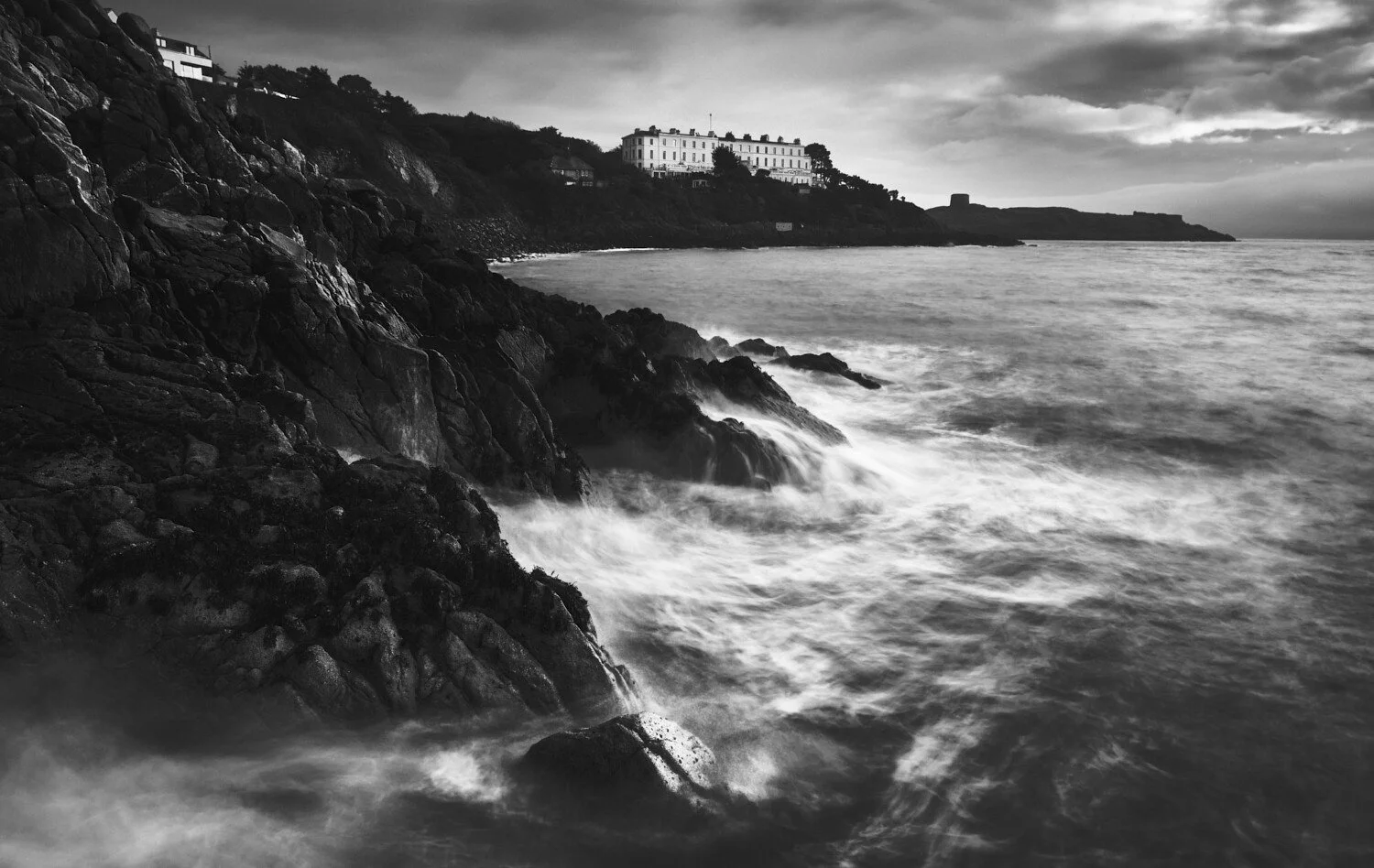 Black and white photo of a rocky coastline with waves crashing against the rocks, a large white building on a hill, and a cloudy sky.
