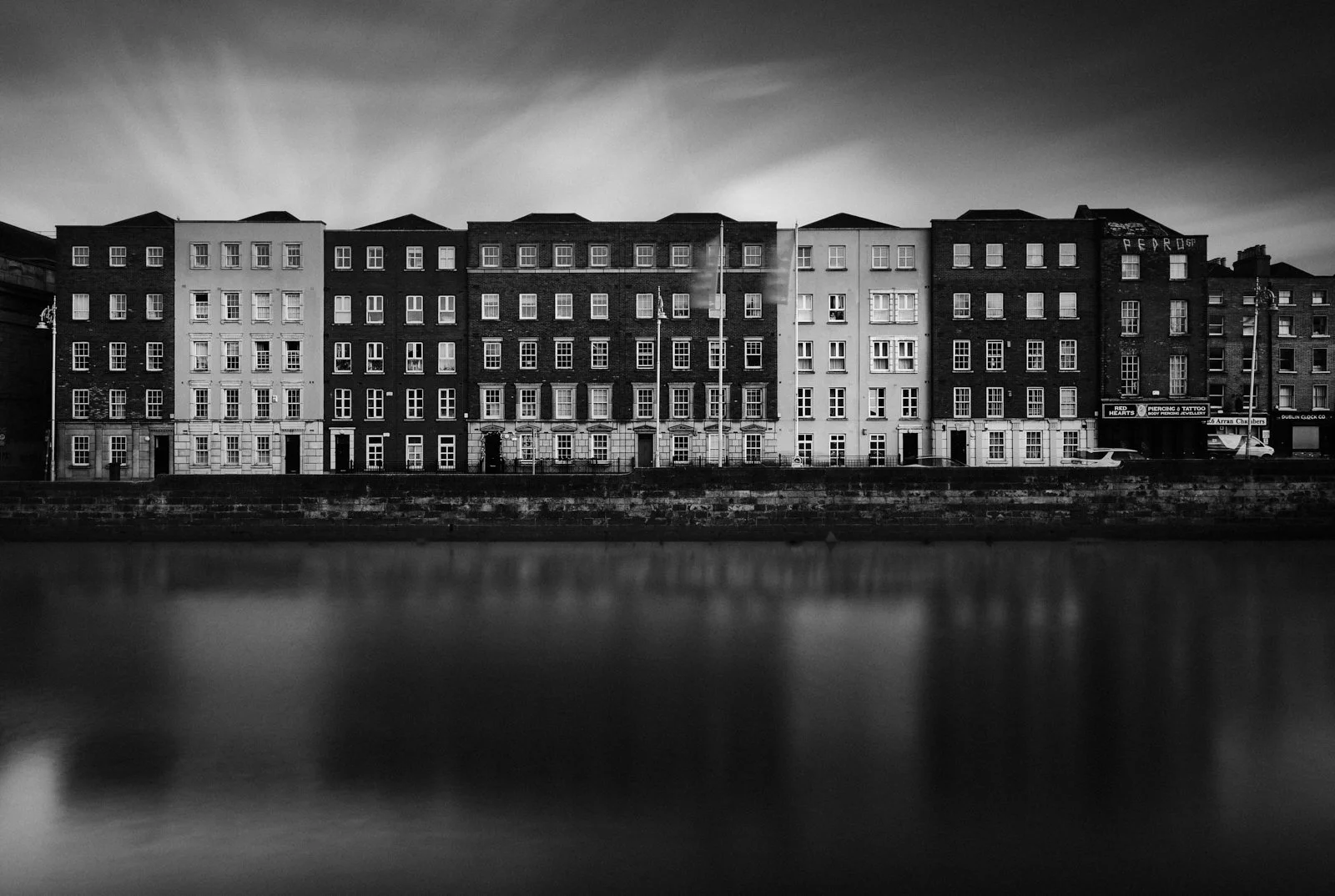 Black and white photo of a row of multi-story buildings along a waterfront, with a cloudy sky overhead.