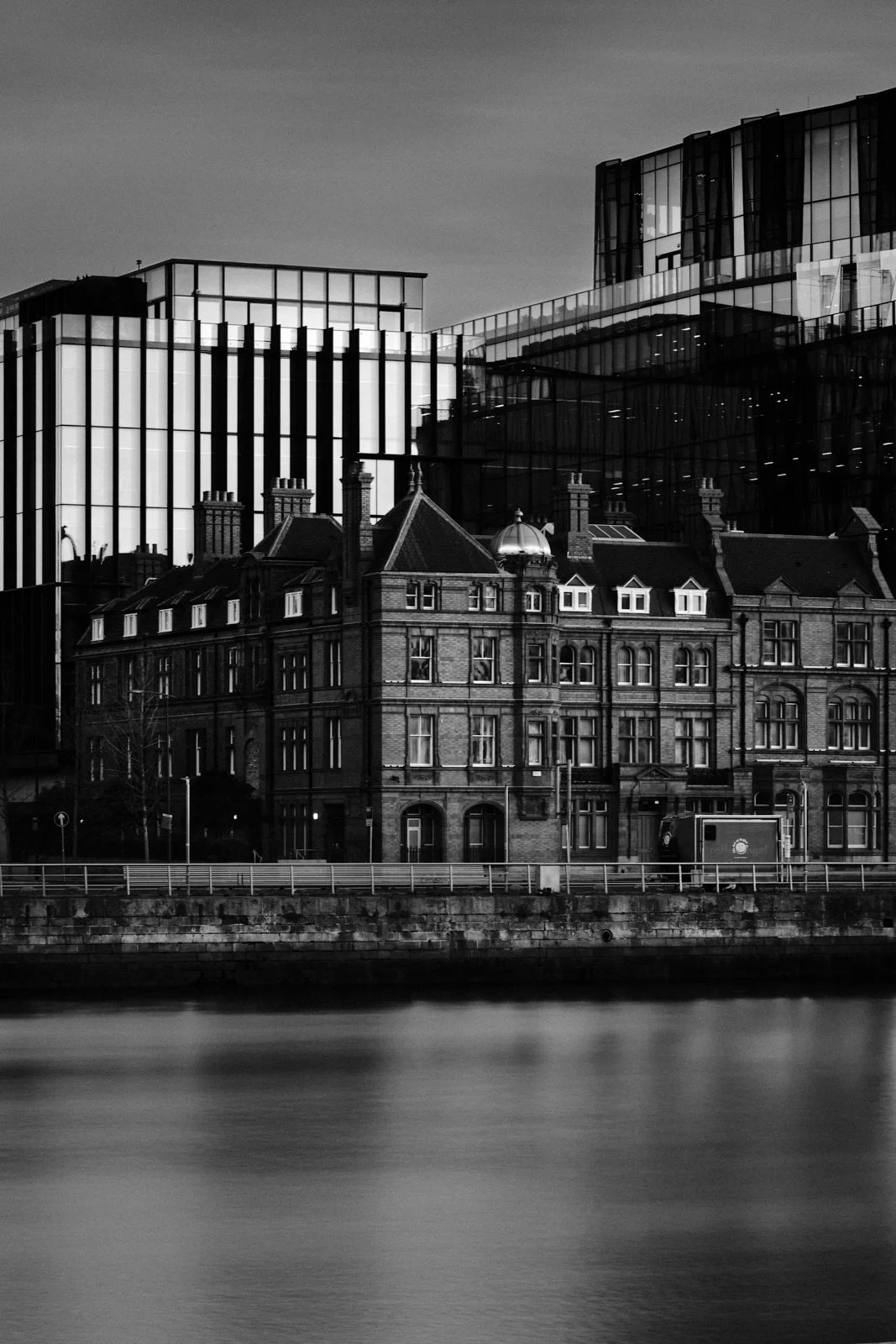 Black and white photo of modern glass buildings behind historic brick buildings by a river.