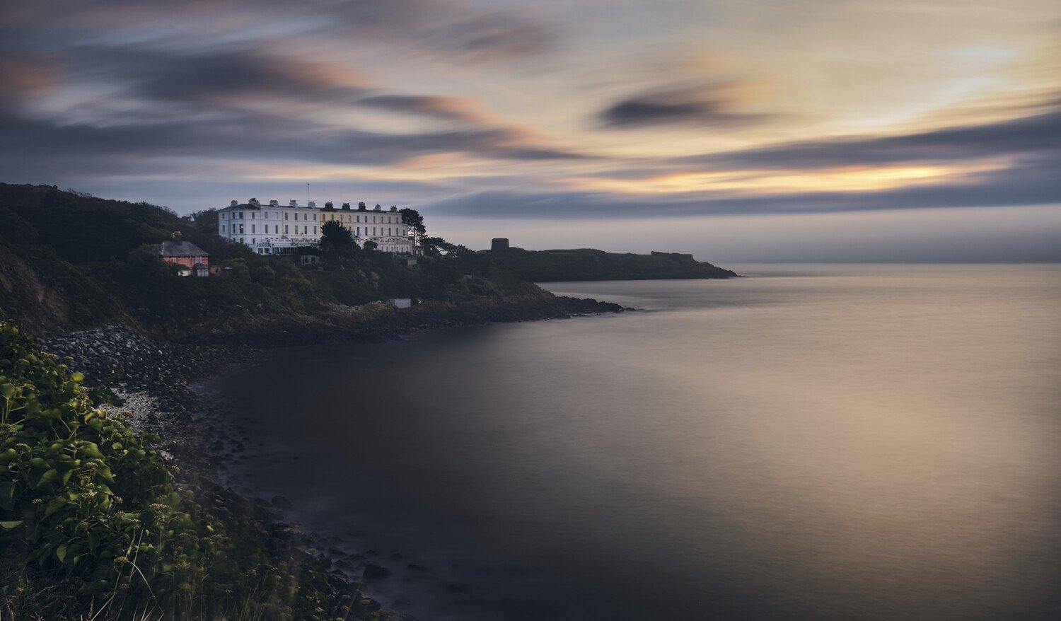A coastal scene at sunset with a white hotel on a cliff, dark rocks and vegetation along the shoreline, and a calm sea with a cloudy sky in the background.